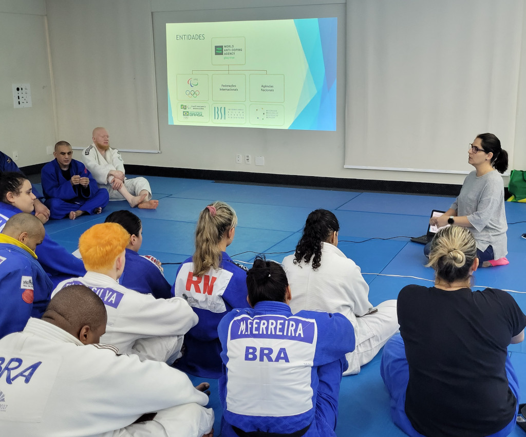 Photo taken in a white room with a blue mat. On the wall a projection with writing in
black with drawings in blue and green. In this projection, it is possible to read the
information about the sports entities related to anti-doping, the highest being WADA
and below the IPC, IBSA and others that are not legible.
There are more than 12 people sitting on the mat in this photo, all but the teacher
being judokas (male and female) in blue and white kimonos. Ahead, the teacher with
brown hair tied back, wearing a gray blouse and glasses talking to the athletes.