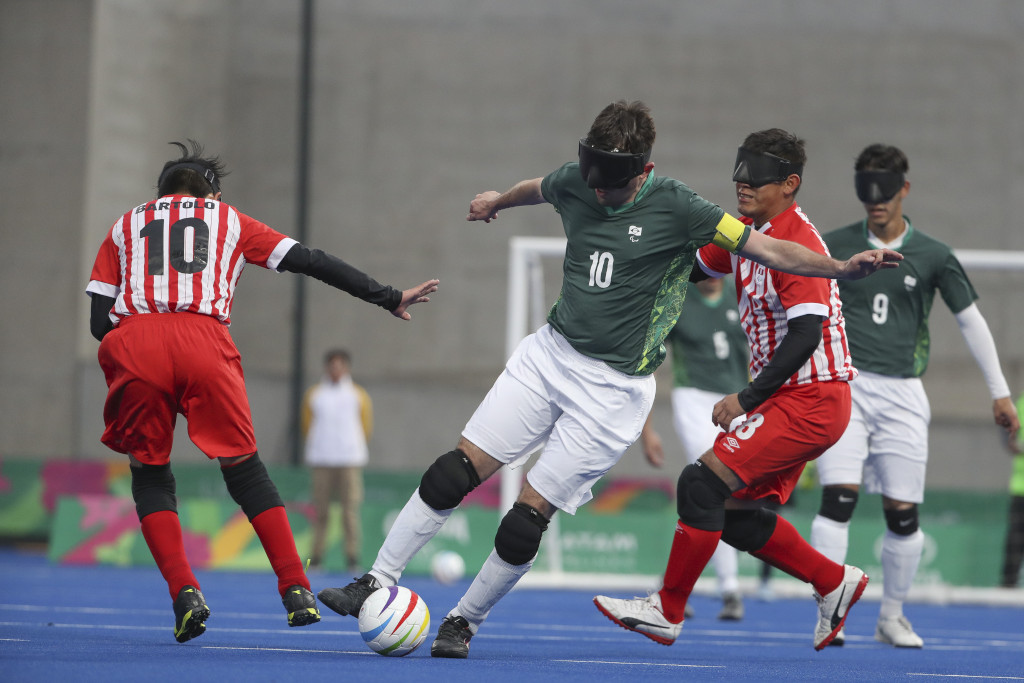 Photo taken during the Lima 2019 ParapanAmerican Games on the blue synthetic
grass field. In the background, a volunteer standing in the uniform of the games, with
brown pants and a white and yellow jacket; the Goal with goalposts and white net and
even more in the background plastic prisms with approximately 60 cm of height with
the arts of the event, in green, pink, orange and white.
In the focus of the photo there are 4 players, 2 athletes from Brazil and 2 from Peru.
The Brazilian players are wearing a green shirt with some green and yellow details on
the sides, shorts and white socks and black knee pads. One of them is wearing the
yellow captain's armband and black cleats (shirt 10) and one is wearing white cleats
(shirt 9).
The athletes from Peru are wearing a black long-sleeved T-shirt underneath the game
shirt, which is striped red and white, with red shorts and socks and black knee pads.
One of them is wearing black cleats (shirt number 10) and one is wearing white cleats
(shirt number 8).
All athletes are wearing the black blind Football mask.