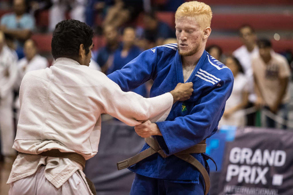 Photo in a gymnasium with several spectators sitting in a red bleacher watching and
cheering for two judokas during the International Grand Prix.
The athlete on the back has brown hair and wears a white kimono and a black belt,
while the athlete in front of him, who is taller than the other athlete, has blond hair, as
he is an albino, and wears a blue kimono, and also has the black belt. Both are holding
the opponent's judogui.