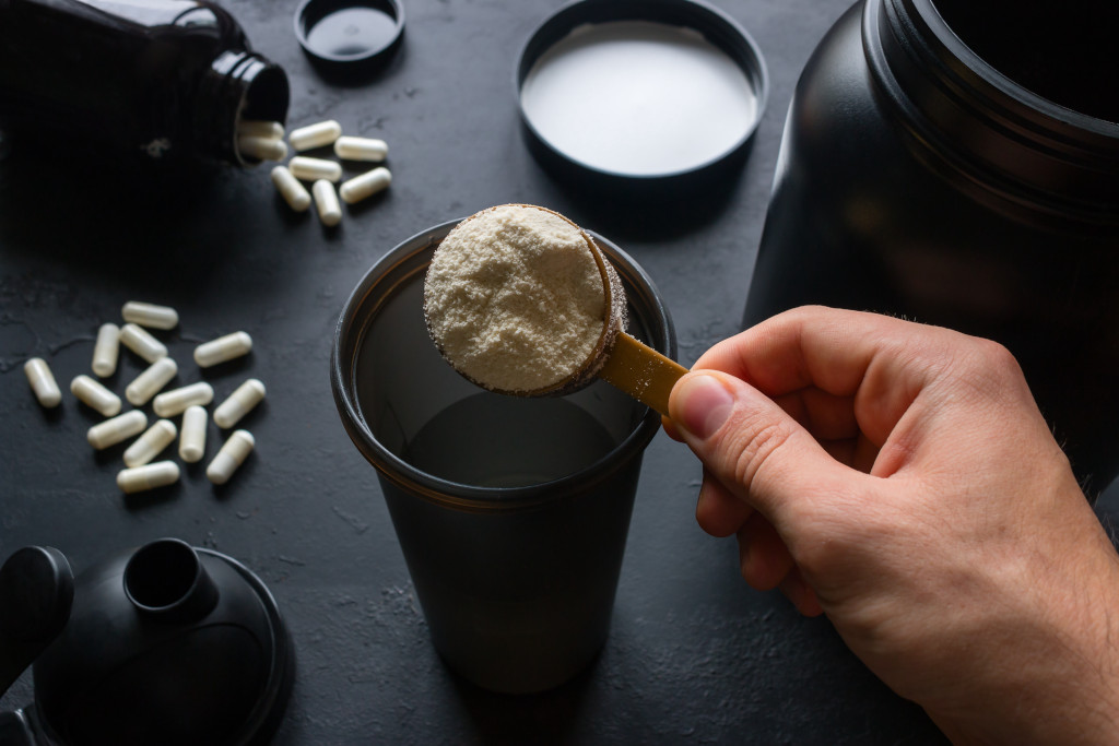 Photo with few colors, the vast majority black and white, of a table with some
supplements, with a black pot with white capsules, other capsules distributed on the
table, plus a large black pot. Also, a man's hand pouring a scoop full of powder, which
appears to be something like whey-protein in a shaker cup.