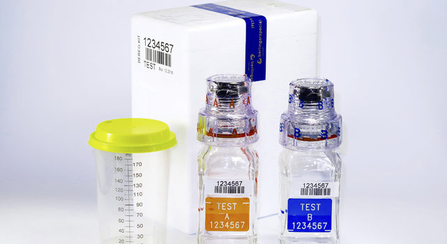 Photo of a Test Kit. Photo taken with a white background showing a white styrofoam
box with a purple ribbon that is the box's seal, and a white label with numbers and a
bar code. The front of the box on the left side has a 180ml clear plastic cup and a lime
green lid with a small opening.
Also in front of the styrofoam box are two clear glass bottles with large lids and a
plastic seal over them. These bottles are taller than the plastic cup but thinner.
On each of these bottles it is possible to see a bar code and an ink marking written
“TEST A, 1234567” in orange, and “TEST B, 1234567” in blue.